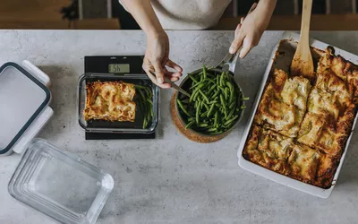 A person using a digital kitchen scale to portion lasagna and green beans.