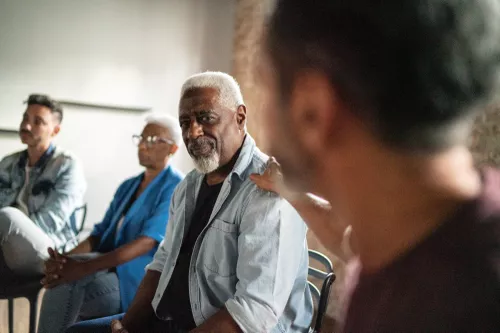 An elderly Black man with a white beard sits in a support group session with a hand on his shoulder.