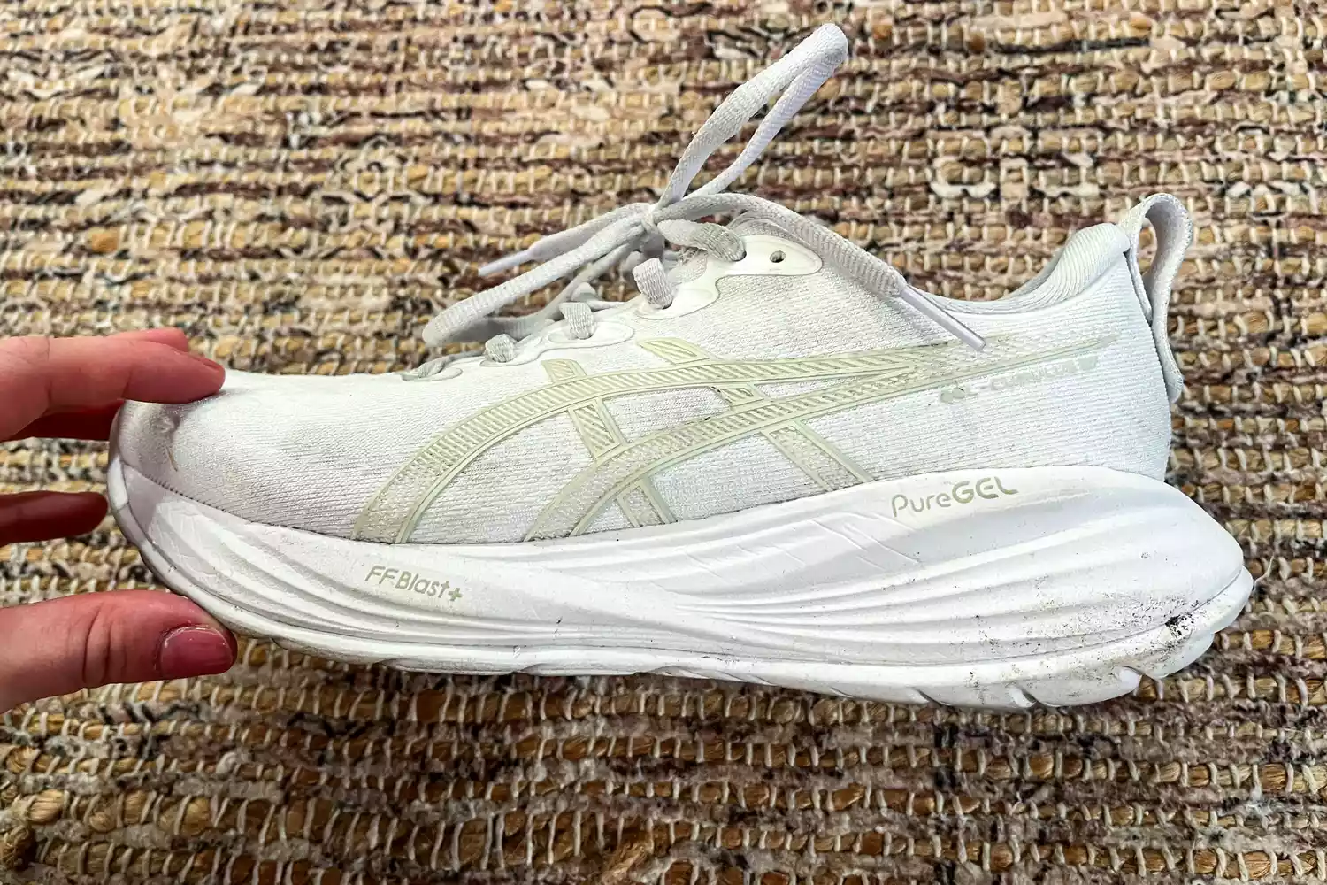 A hand holding a white ASICS Gel-Cumulus 27 shoe, focusing on the PureGEL and FF Blast+ cushioning in the midsole.