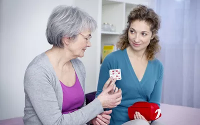 An elderly woman receiving medication guidance from a caregiver at home.