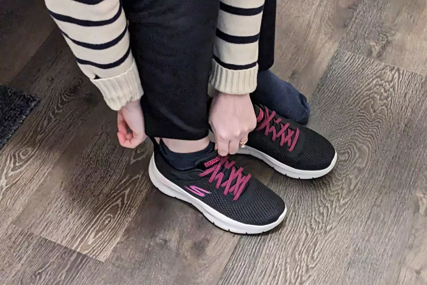 A person tying the laces of black and pink Skechers walking shoes on a wooden floor.