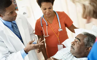 A doctor and a nurse in a hospital room discussing a care plan with an elderly male patient in a hospital bed.
