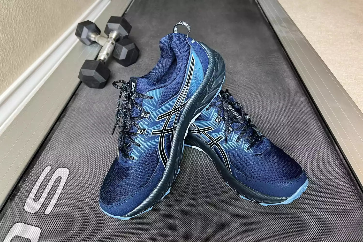 Blue running shoes on a treadmill belt next to dumbbells in a gym setting.