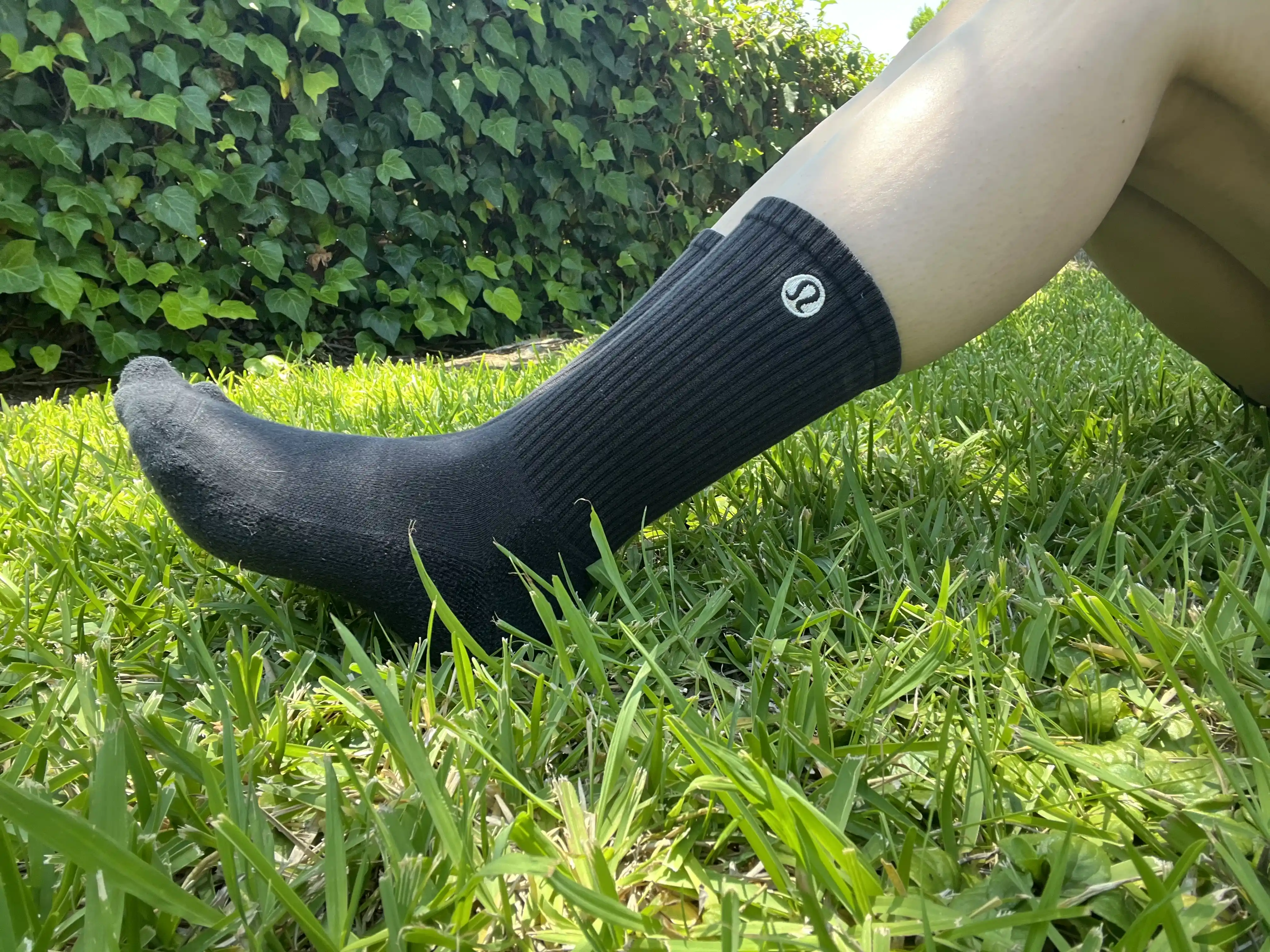 A person resting their leg on green grass while wearing black Lululemon crew socks.