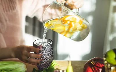 Fresh lemon-infused water being poured into a glass on a kitchen counter with fresh produce.