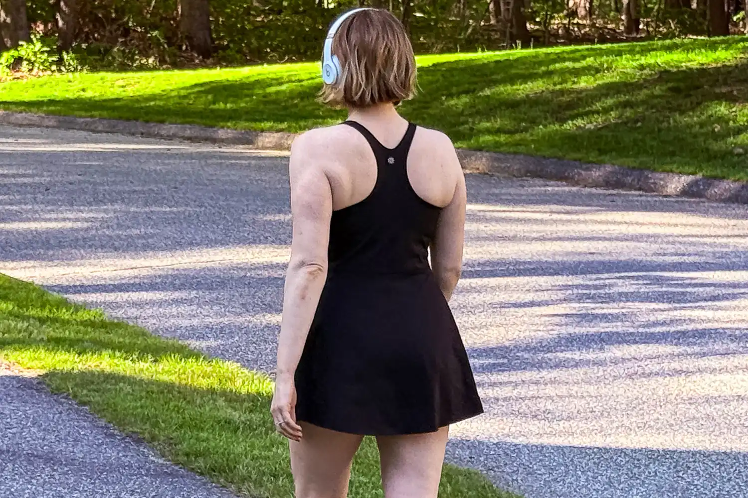 A woman wearing a black athletic dress and headphones walks on a paved park path.