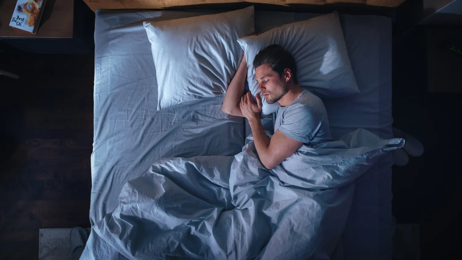 A man sleeping peacefully on his side in a dimly lit bedroom with blue sheets.