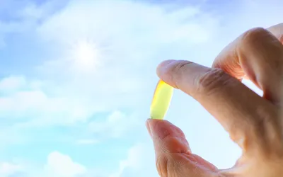 A hand holding a yellow Vitamin D softgel against a bright sun and blue sky.