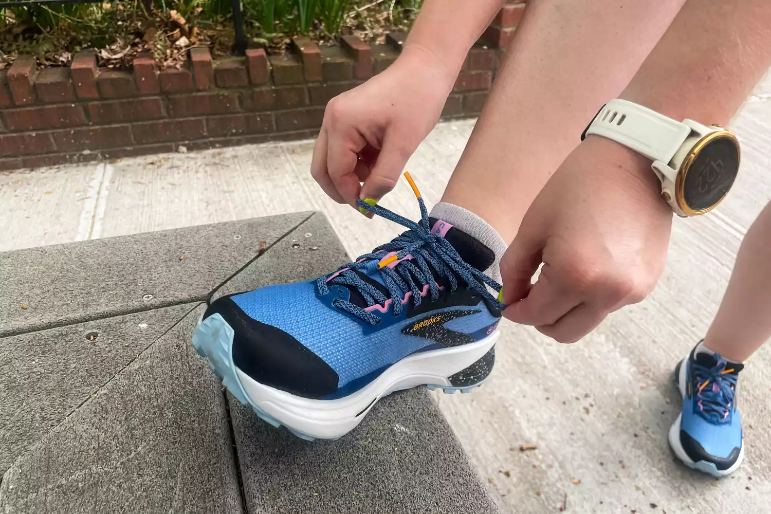 A person tying the laces of a trail running shoe while wearing a fitness watch.