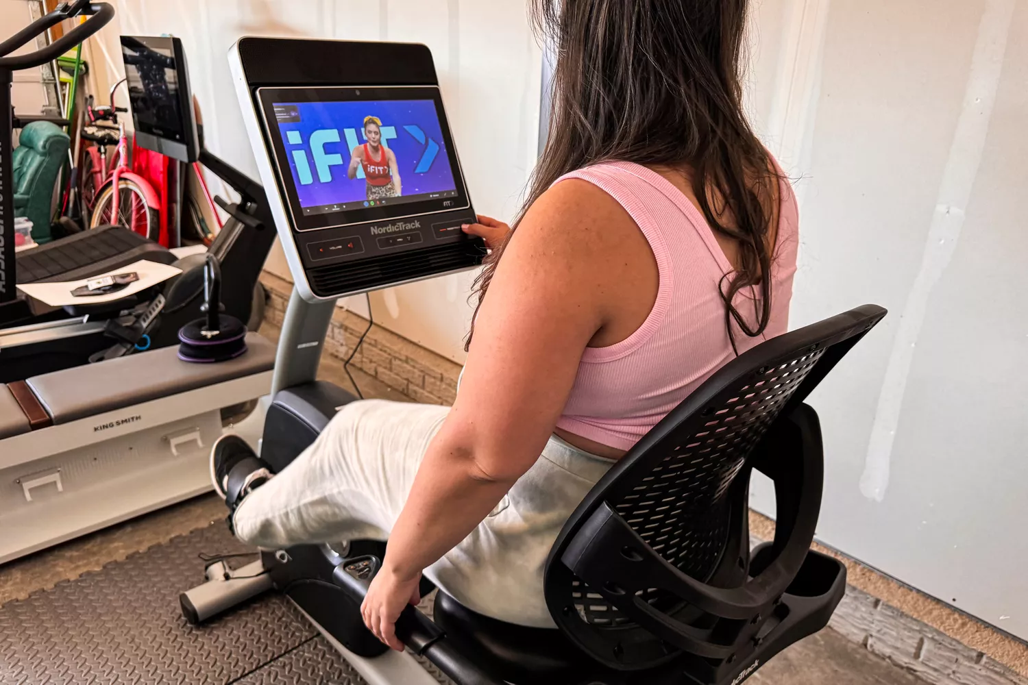 A woman using the touchscreen display on a NordicTrack recumbent bike with an iFIT trainer visible.