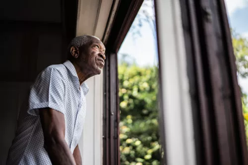 An elderly man in a patterned shirt stands by a window, gazing outside.