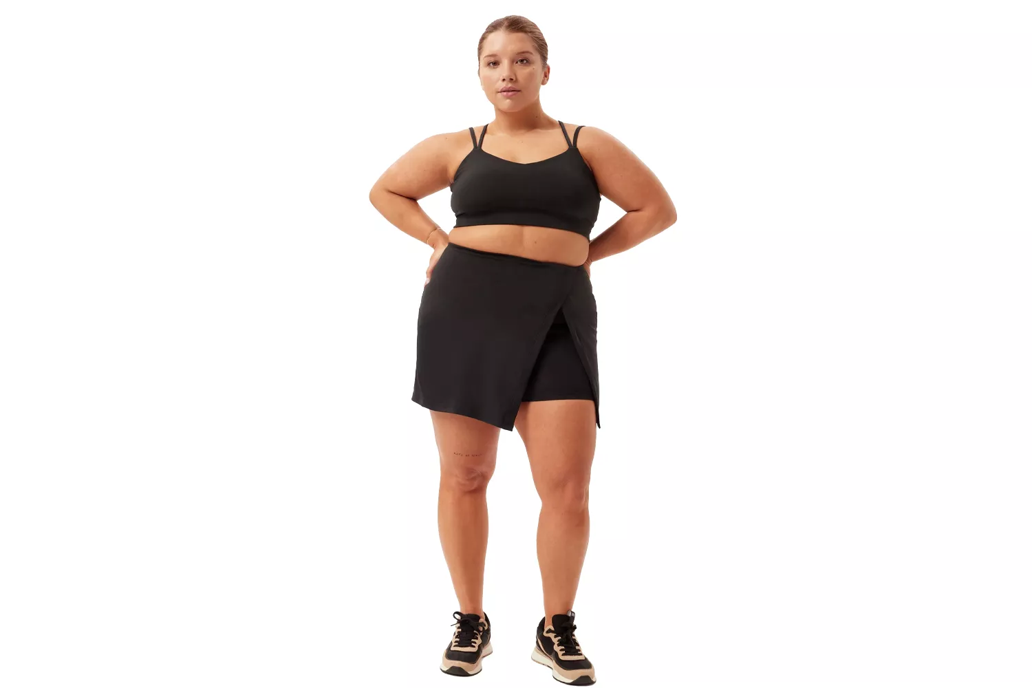 A plus-size model wearing a black athletic skort and sports bra against a white background.
