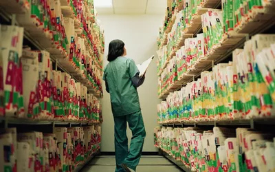 A healthcare worker in scrubs walking through an aisle of medical records and patient files.