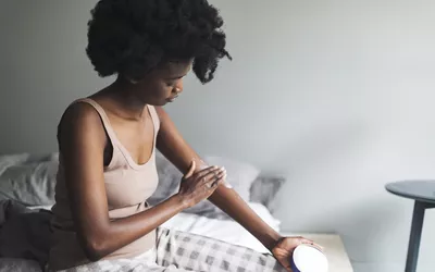A woman sitting on a bed applying lotion to her arm.
