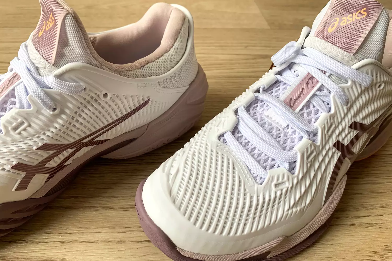 Close-up of white and rose gold Asics Court FF 3 shoes on a wooden surface.
