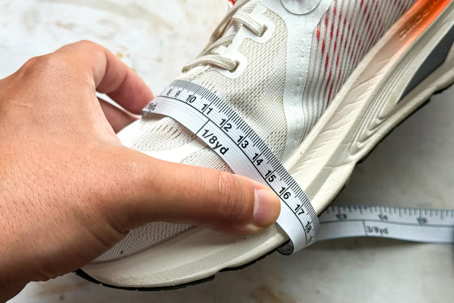 A hand using a measuring tape to measure the length and width of a white athletic sneaker.