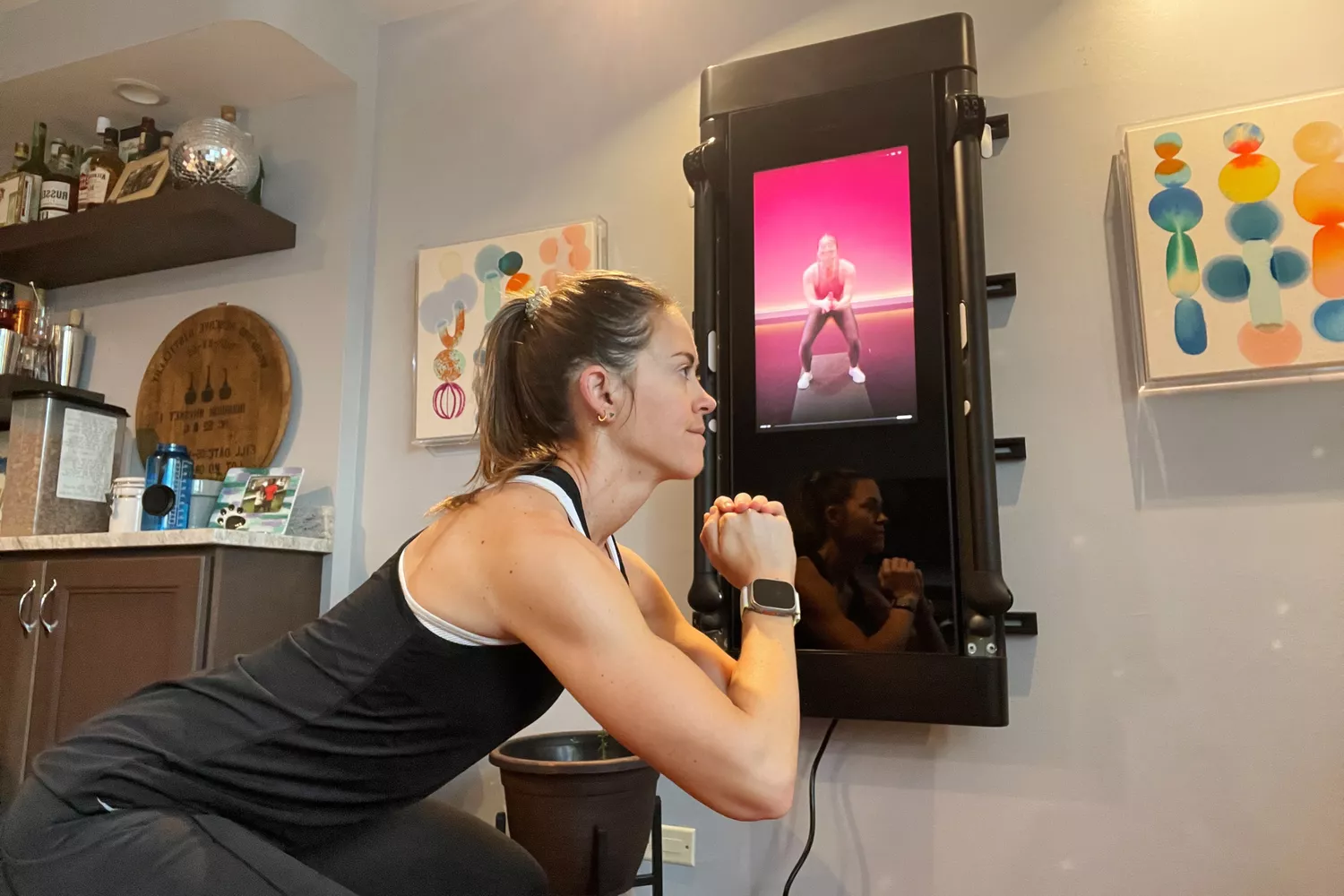 A woman performing squats in front of a wall-mounted Tonal smart fitness system.