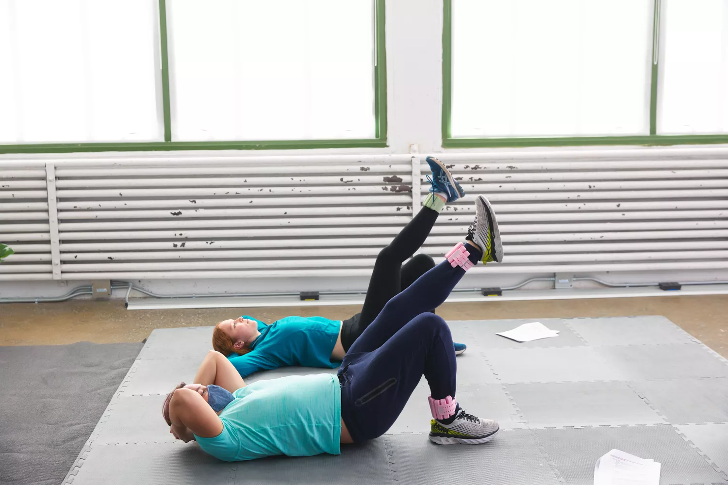 Two women performing leg raises and planks with ankle weights in a gym.