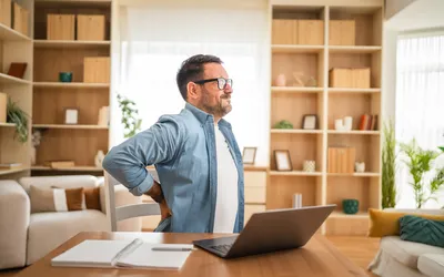 A man in a home office holding his lower back in discomfort while sitting at a desk.