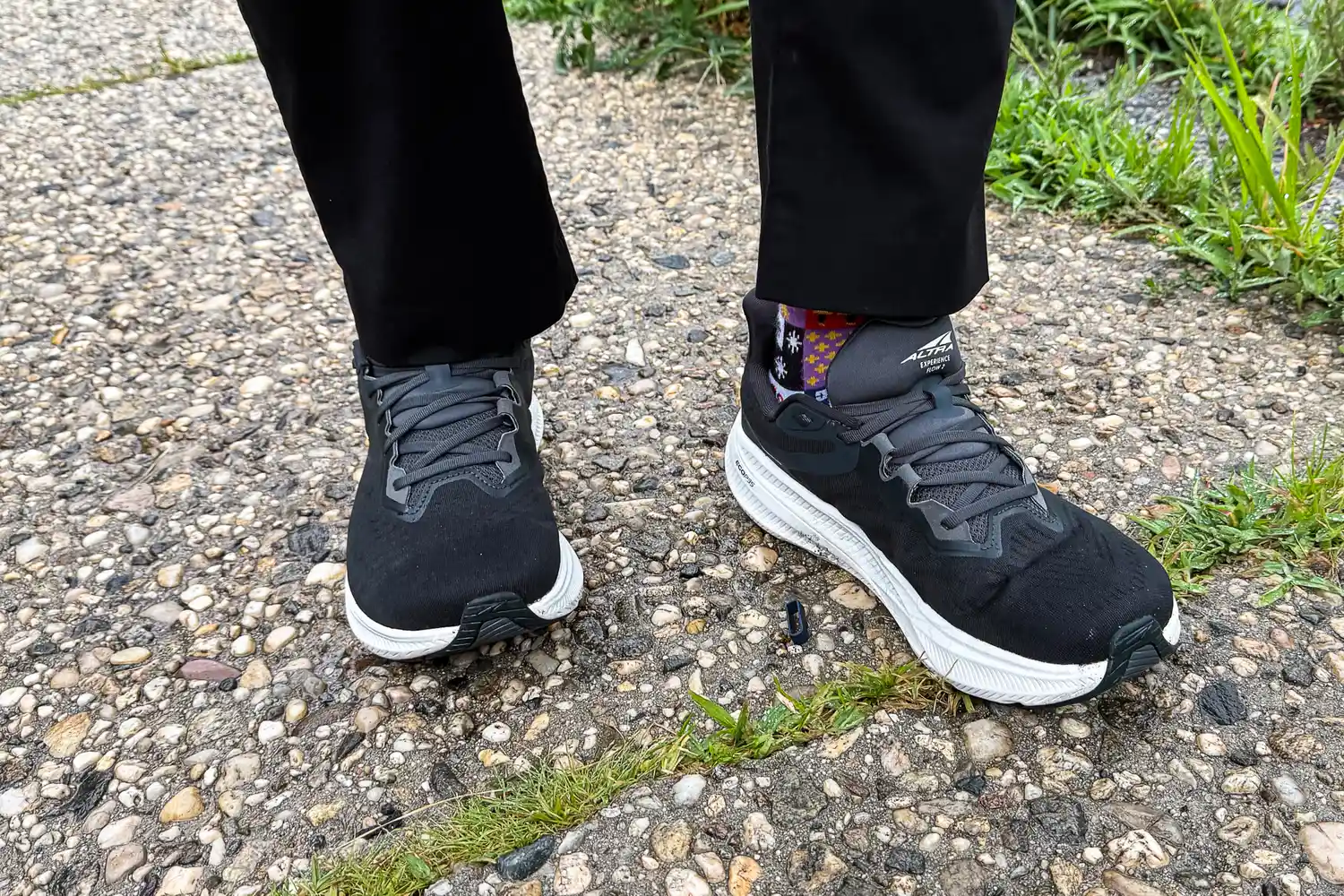 A person wearing black Altra Experience Flow 2 shoes with colorful socks standing on a pebble-covered path.