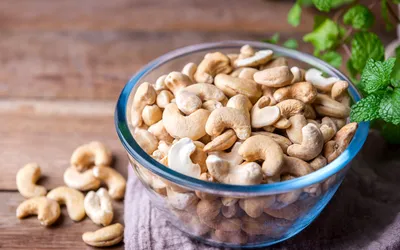 A glass bowl of raw cashew nuts on a wooden table with fresh mint.