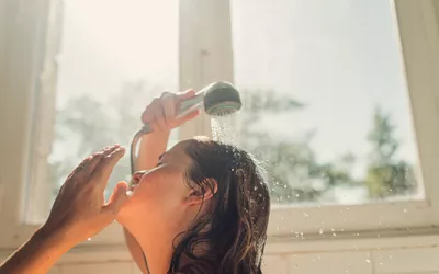 A person washing their hair and body under a showerhead with splashing water.