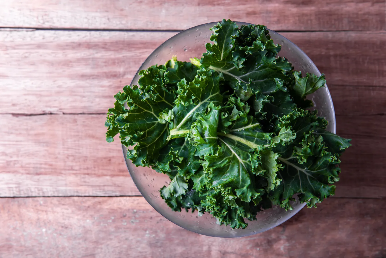 A clear bowl filled with fresh green kale placed on a rustic wooden table.