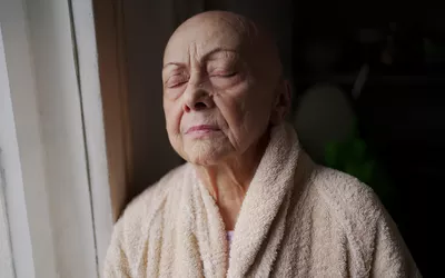 An elderly woman looking out a window in a quiet, sunlit room.