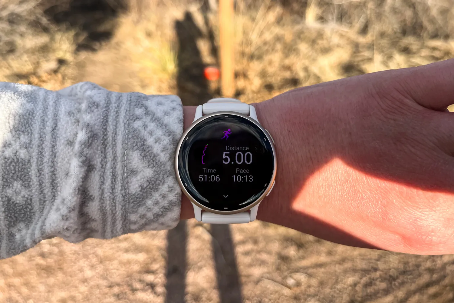 A white smartwatch displaying running distance, time, and pace metrics during an outdoor trail run.