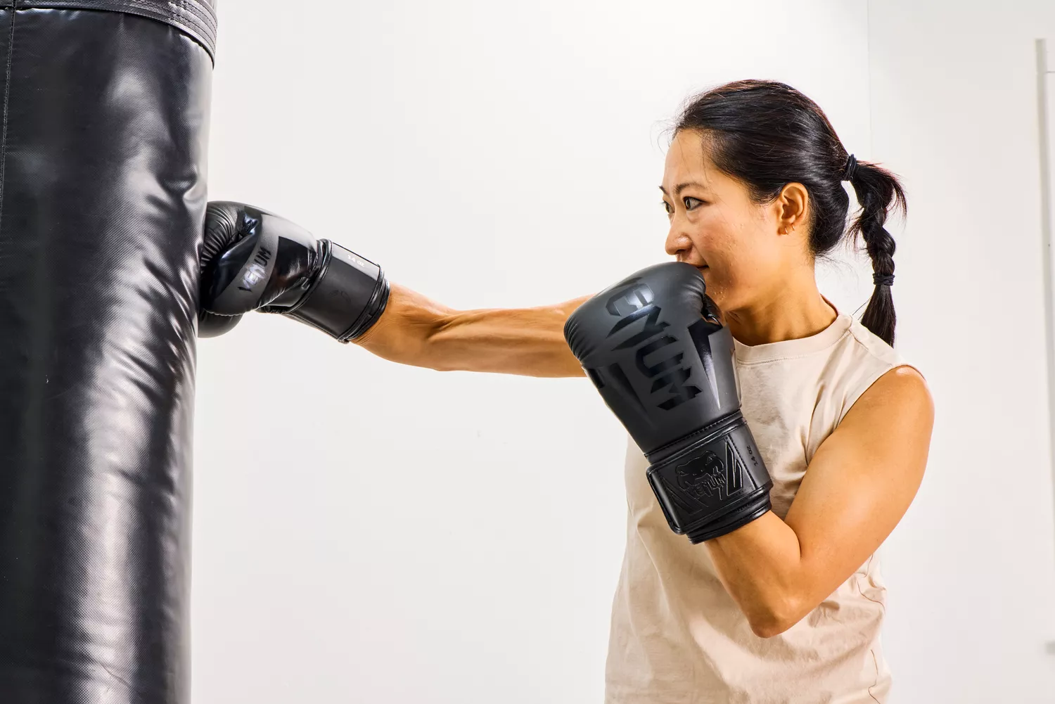 A woman in a beige tank top using black Venum gloves on a heavy bag.
