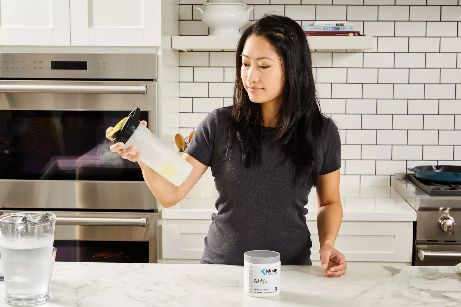 A woman in a kitchen preparing a nutritional shake with a scoop of white powder.
