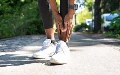 A person in athletic gear holding their calf in pain while outdoors.
