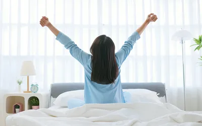 A woman in a light blue shirt stretching her arms upward in a bright, white bedroom.