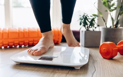 A person standing on a digital scale in a room with a foam roller and dumbbell.