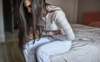 A woman sitting on a bed holding her stomach in discomfort.