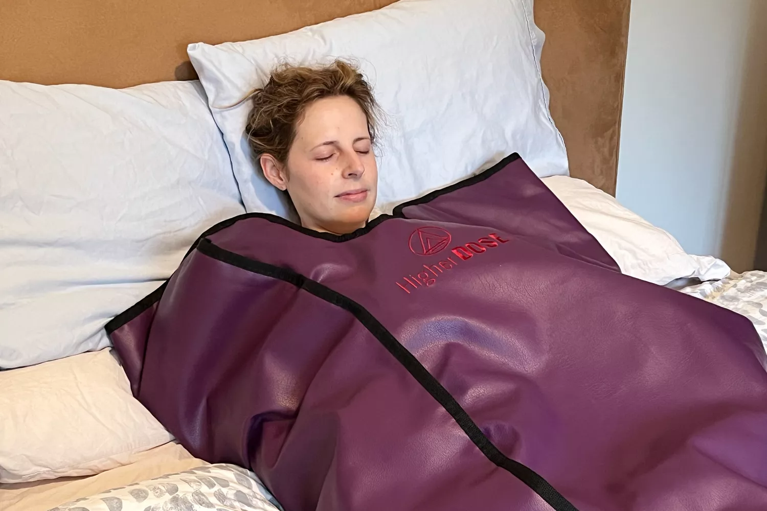 A woman using a black HigherDOSE infrared sauna blanket while lying on a bed.