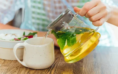 A glass teapot pouring mint tea into a mug next to a healthy meal, suggesting a moment of peace.