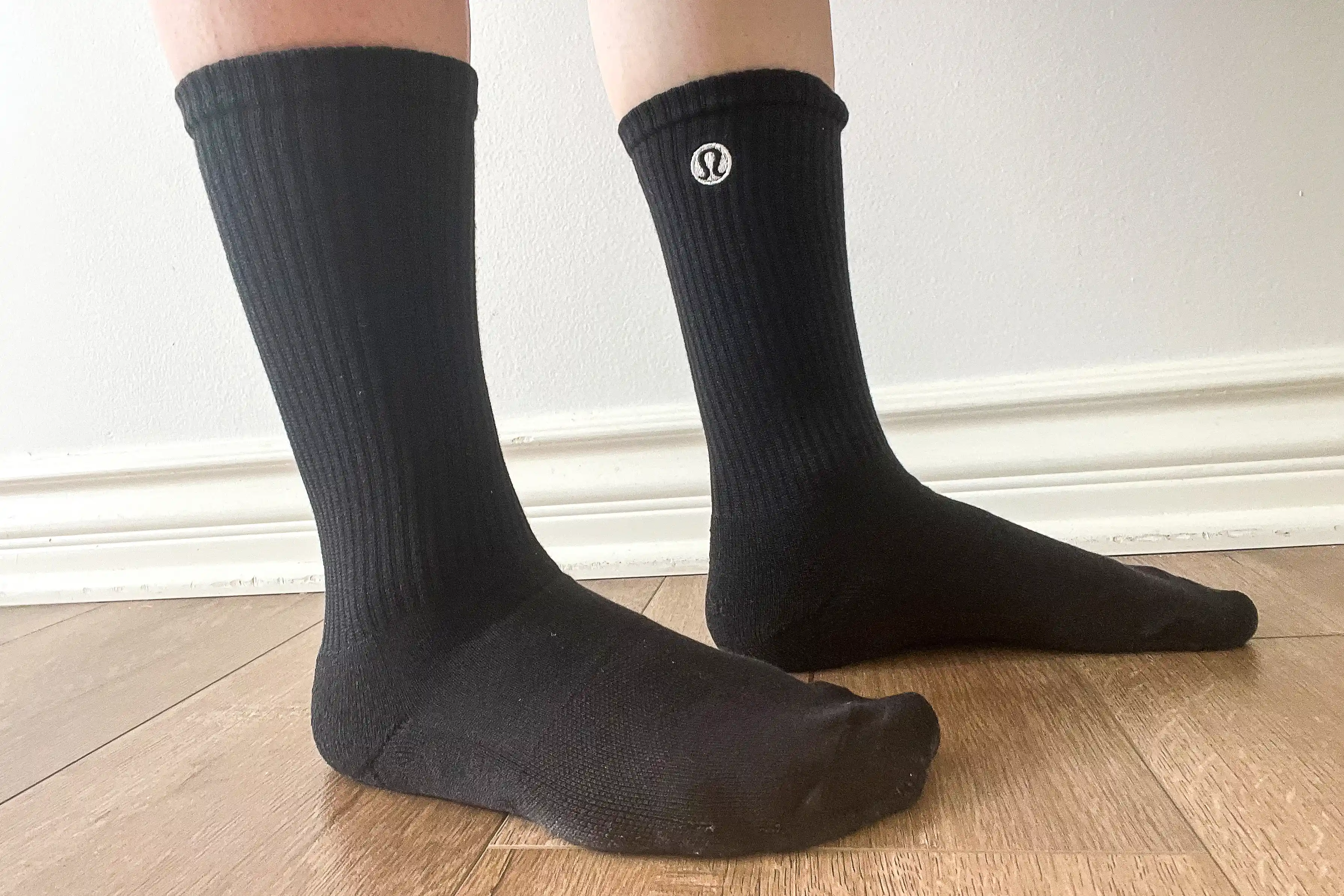 A person wearing black Lululemon crew socks standing on a wooden floor, highlighting the arch support and snug fit.