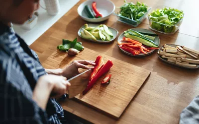 A person slicing red bell peppers on a wooden board surrounded by fresh vegetables.