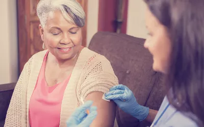 A healthcare worker in blue gloves administering an injection to an elderly patient at home.