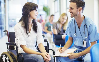 A medical provider in blue scrubs talking to a patient in a wheelchair in a clinical hallway.