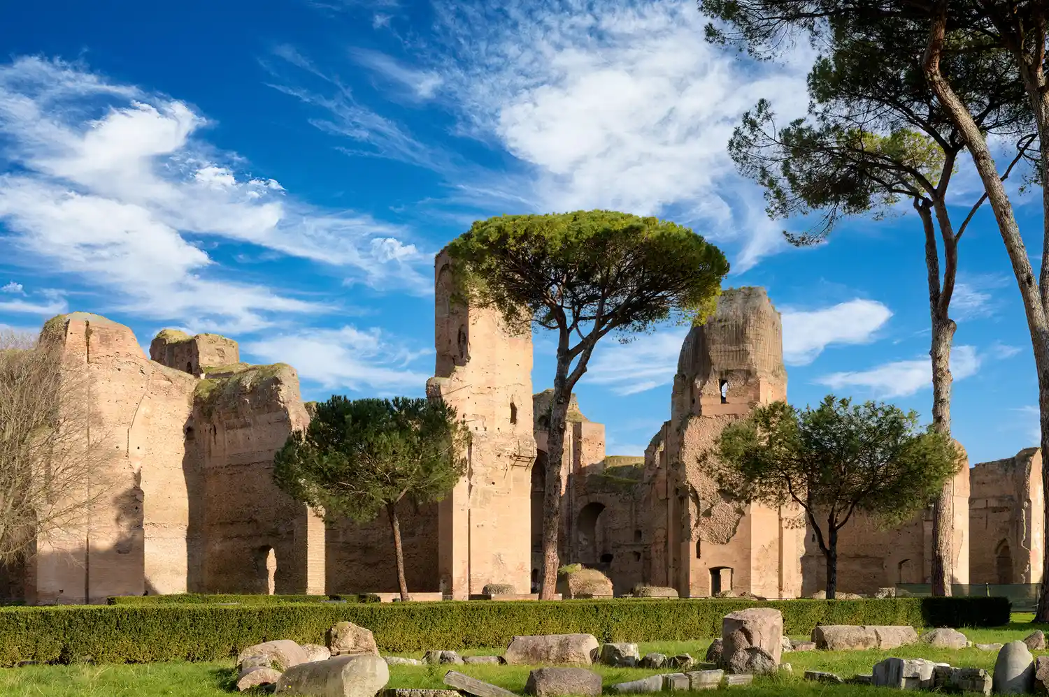 The towering brick ruins of the Baths of Caracalla set against a clear blue sky and green grass.