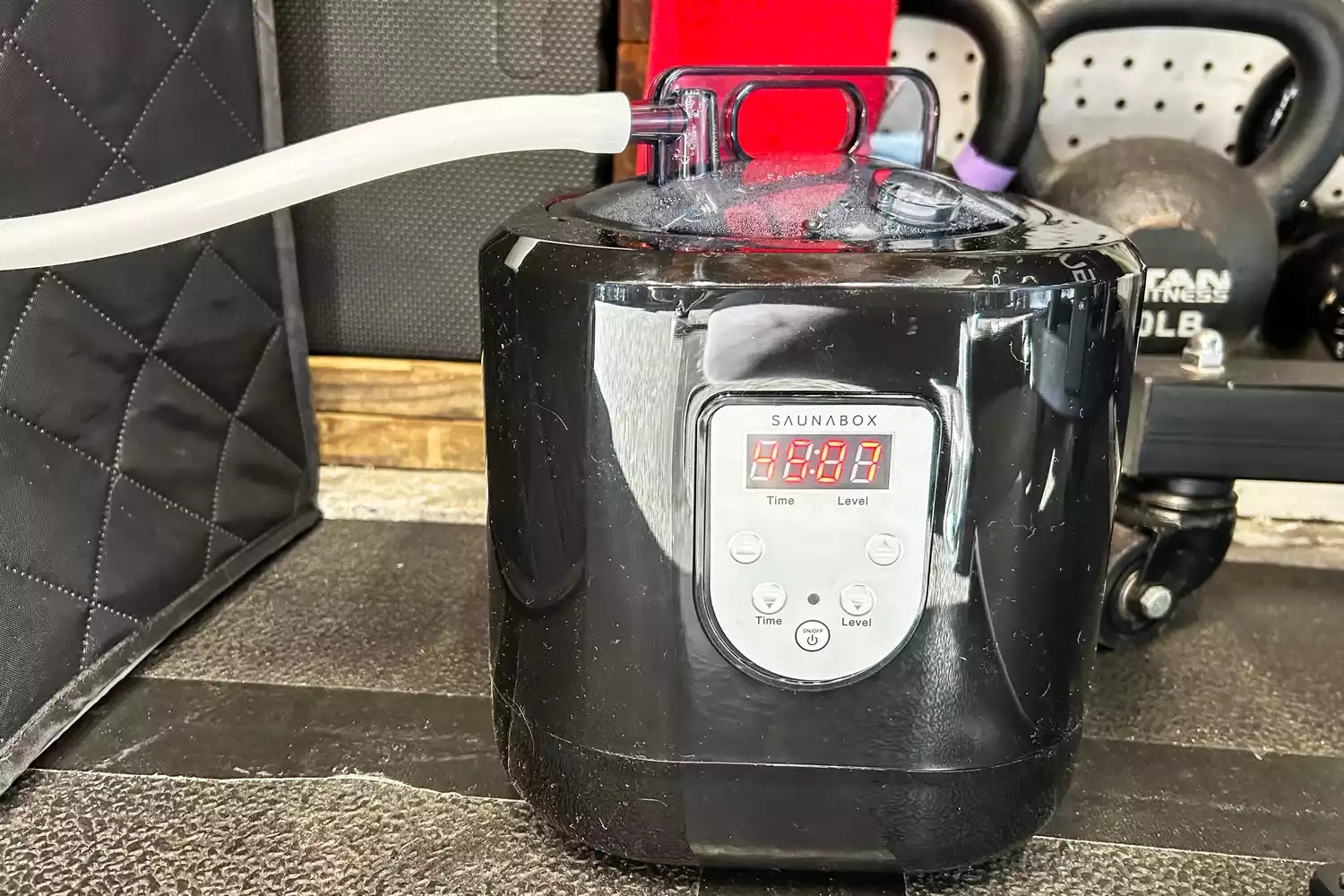 A SaunaBox SmartSteam generator with a digital display sitting on a gym floor next to a kettlebell.