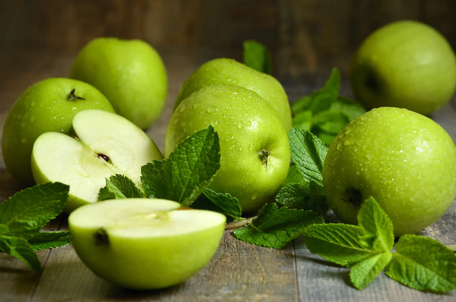 Fresh green apples and mint leaves with water droplets on a wooden background.