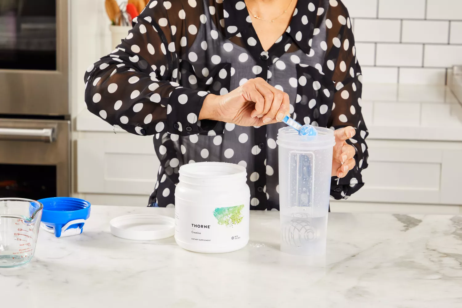 A woman in a polka-dot shirt adding a scoop of supplement powder to a shaker bottle.