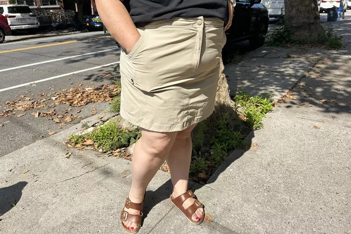 A woman walking on a city sidewalk wearing a beige skirt and sandals.