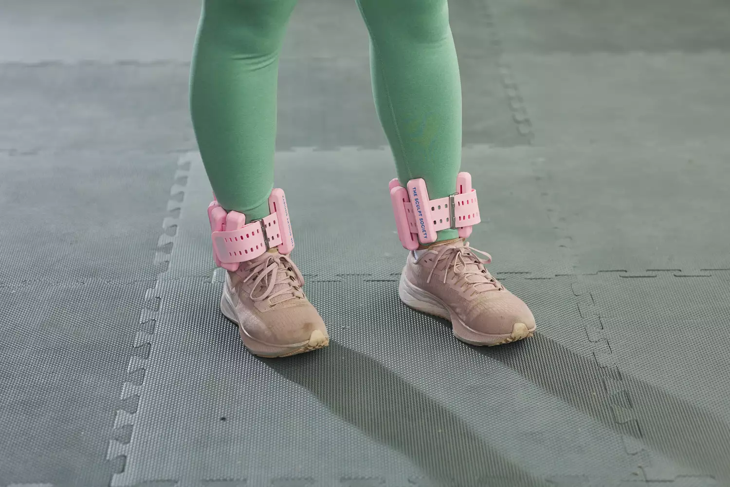 A person standing on a foam mat wearing pink Sculpt Society ankle weights.