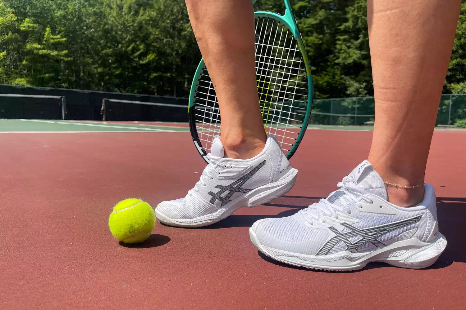 Close-up of Asics tennis shoes on a red clay court next to a tennis ball and racket.