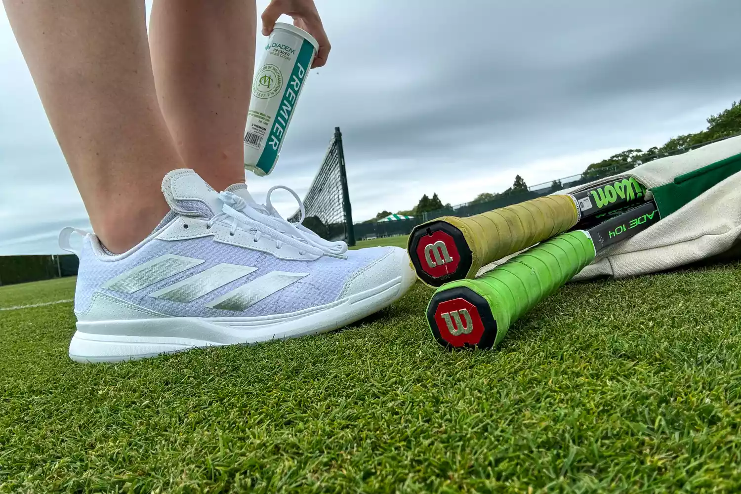 White Adidas tennis shoes worn on a grass court alongside Wilson rackets.