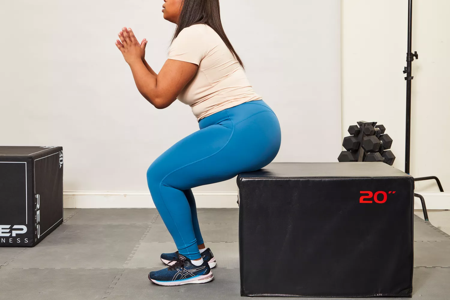 A woman in blue leggings performing a box squat on a 20-inch plyo box in a home gym.